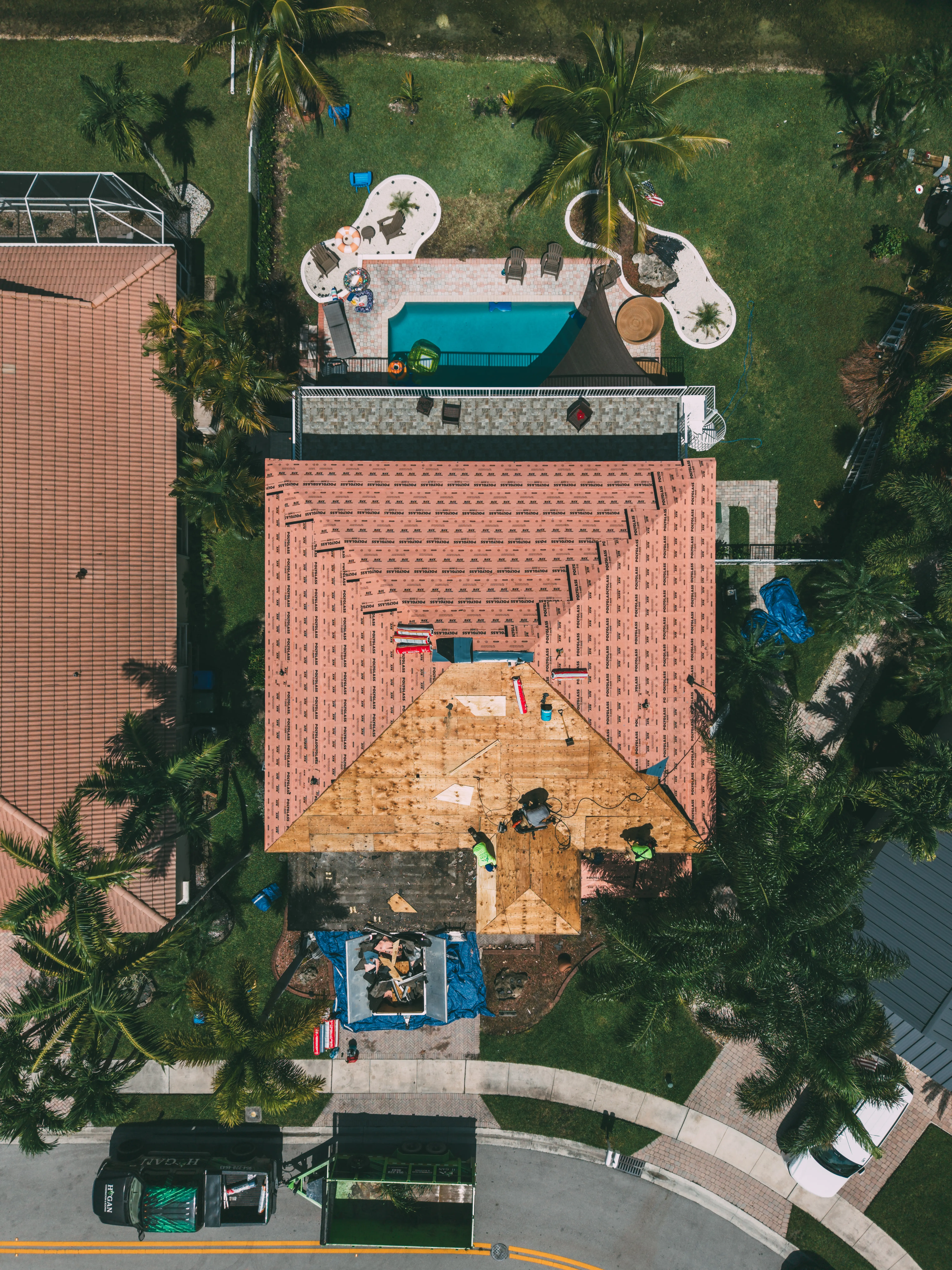 Aerial view of a house undergoing roof replacement, with workers on a partially removed orange tile roof and a backyard pool.